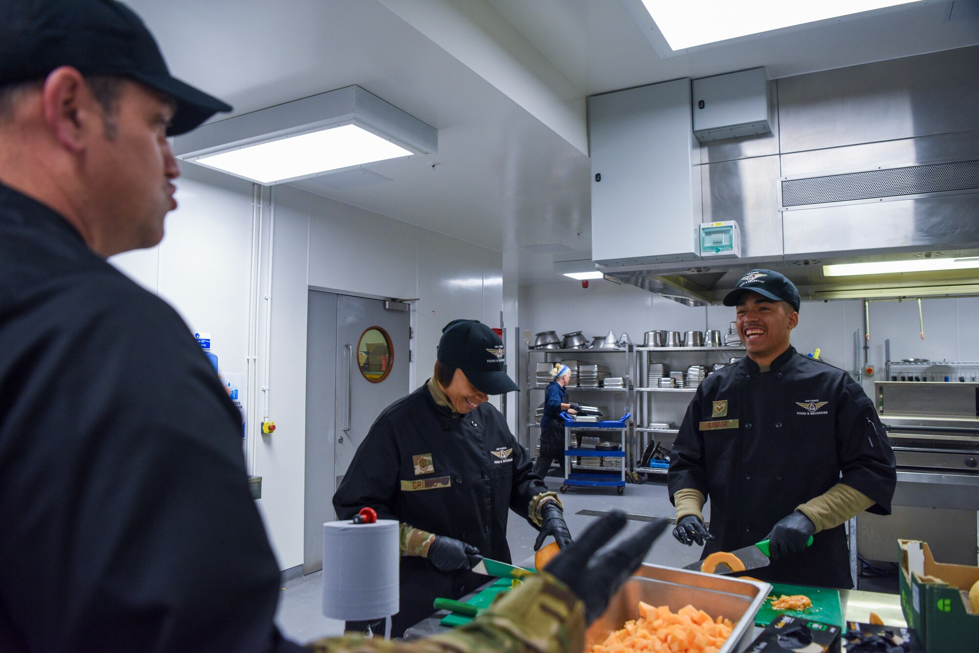 U.S. Air Force Col. Joseph Wall, left, 100th Air Refueling Wing deputy commander, and Chief Master Tiffany Griego, center, 100th ARW command chief, talk while cutting cantaloupe in preparation for a meal with Senior Airman Jurell Savage, 100th Force Support Squadron Gateway Dining Facility storeroom manager, during a “Day in the Life” immersion at Royal Air Force Mildenhall, England, Sept. 13, 2024.