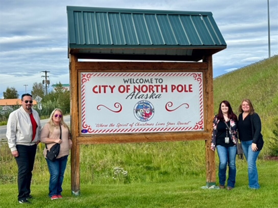 A large sign welcoming people to North Pole, Alaska have four people stand next to it. two on each side. the grass is green with no snow.