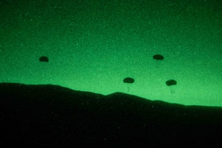 Silhouettes of parachutists in the sky and a mountain range are seen against a green sky in a night vision photo.