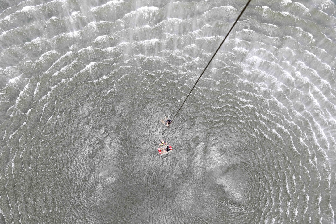 A service member swims near a Coast Guardsman helping a soldier in rescue basket in body of water while a rope hangs from above.