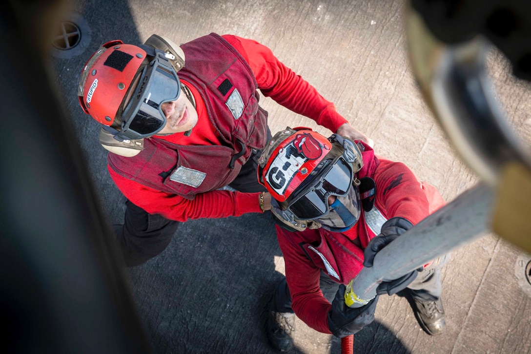 Sailors wearing helmets and goggles connect a rope to a partially visible helicopter as seen from above.