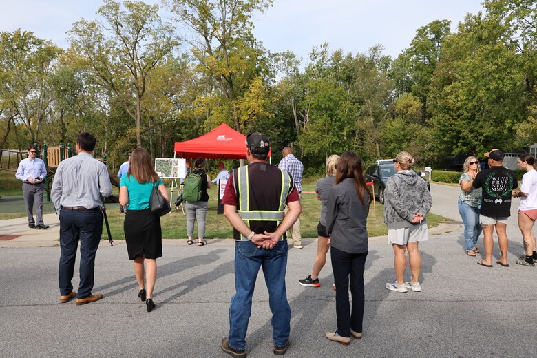 Group stands on sidewalk facing a red canopy in the background waiting for event to begin