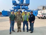 A group of military and civilian personnel pose in front of a crane