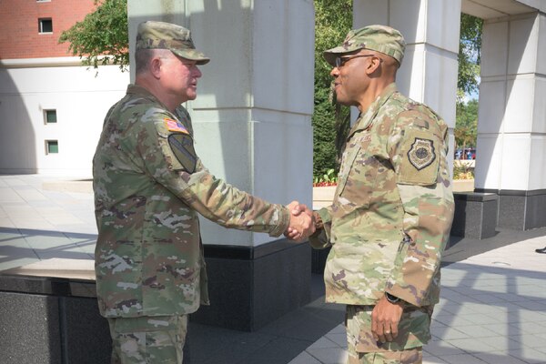 Two service members in uniform shake hands in front of a building.