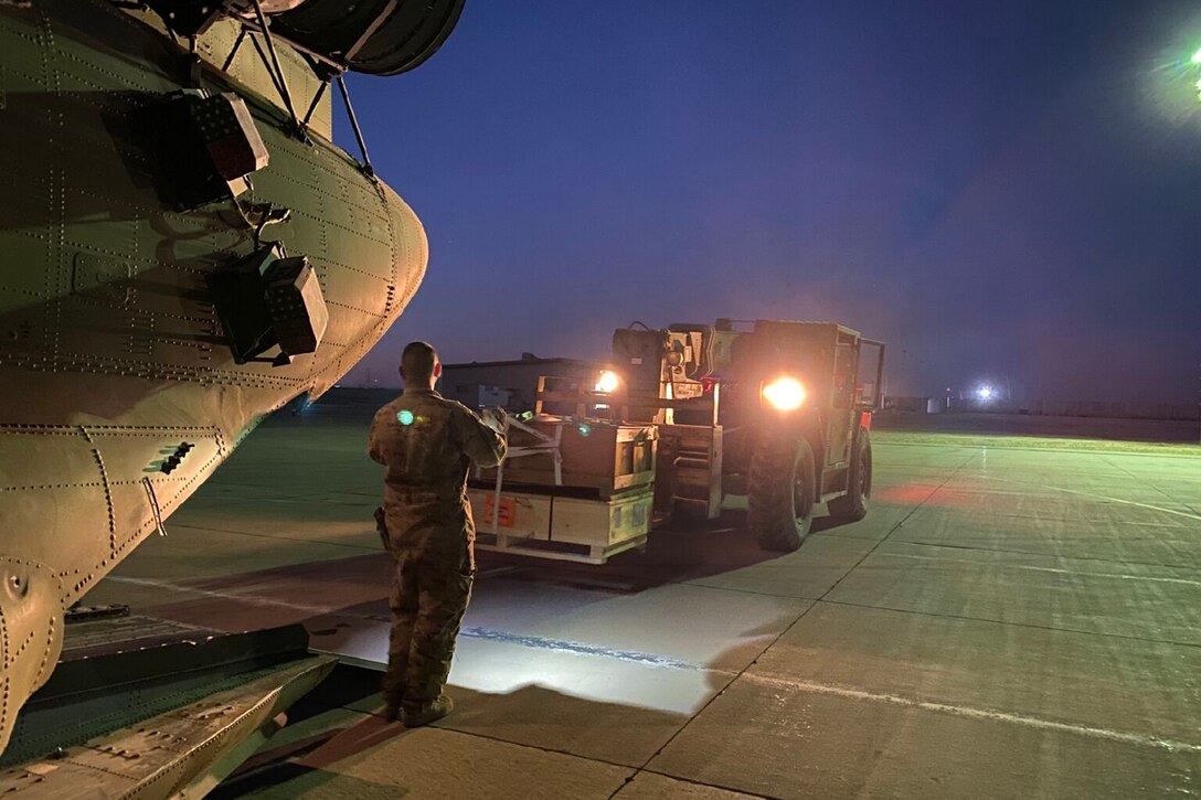 A soldier directs a forklift driver carrying cargo to load it on a helicopter.