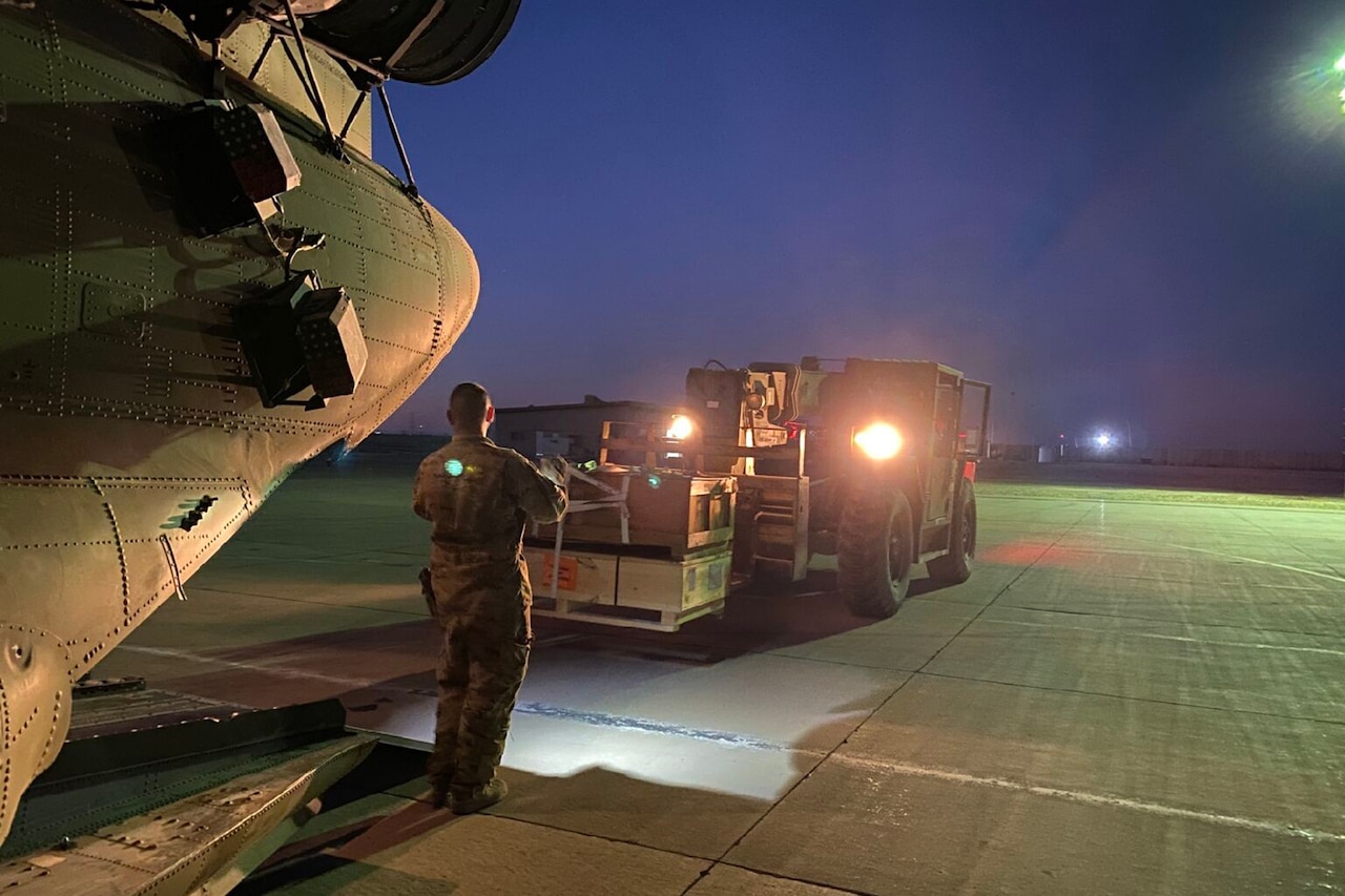 A soldier directs a forklift driver carrying cargo to load it on a helicopter.