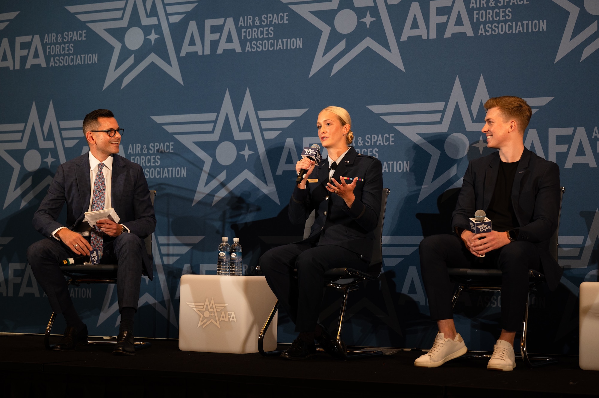 woman in uniform sits on stage and speaks into microphone while two men in civilian clothes sit on either side listening