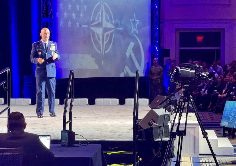 A man in a dress military uniform stands on a stage speaking to a crowd.