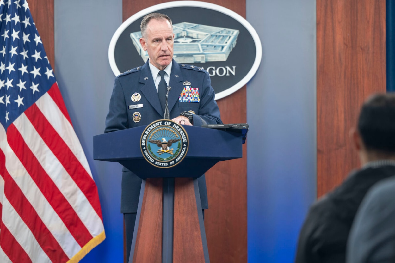 A man in a military uniform stands behind a lectern. Behind him is an American flag. The medallion on the lectern indicates that he is at the Department of Defense.