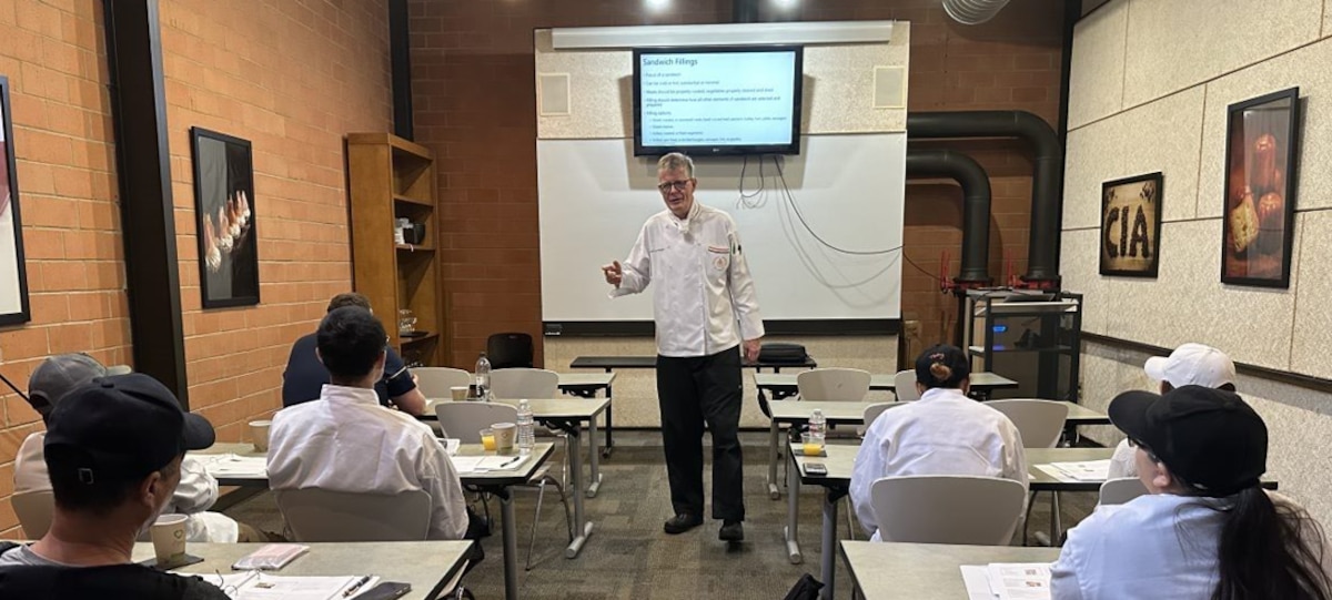 chef stands at front of classroom with two rows of students looking on
