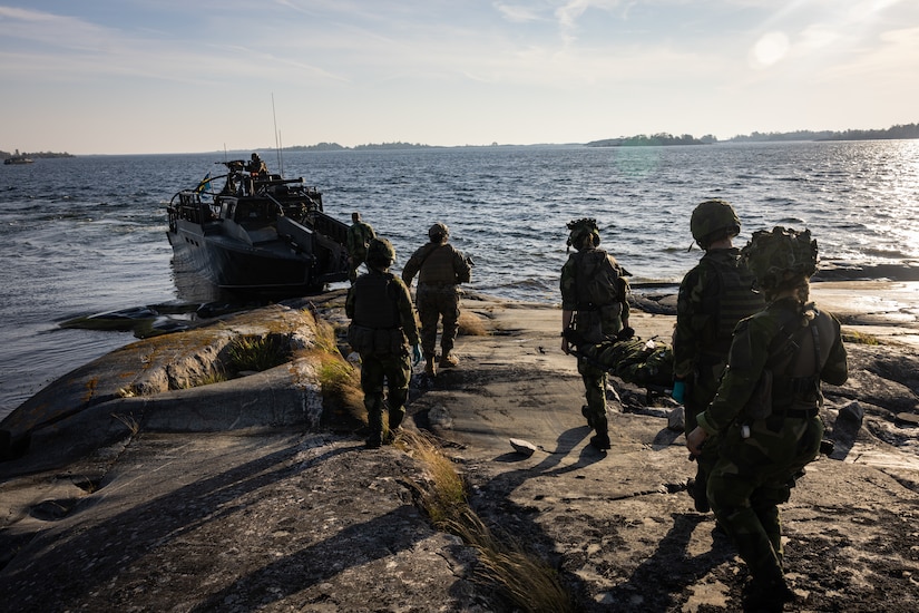 Troops on a land approach an amphibious vehicle emerging from the water.