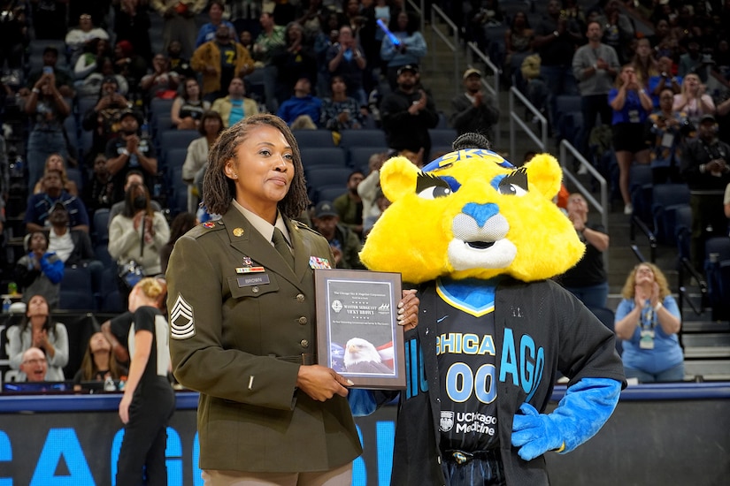 A soldier stands on a basketball court smiling and holding a plaque while posing for pictures with a team mascot dressed as a lioness.