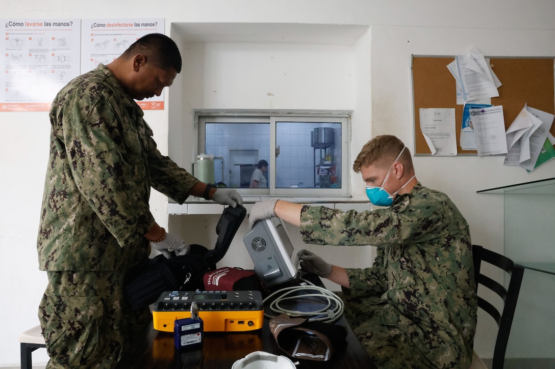 Two sailors, one sitting and the other standing, focus on equipment sitting on a wooden table between them.