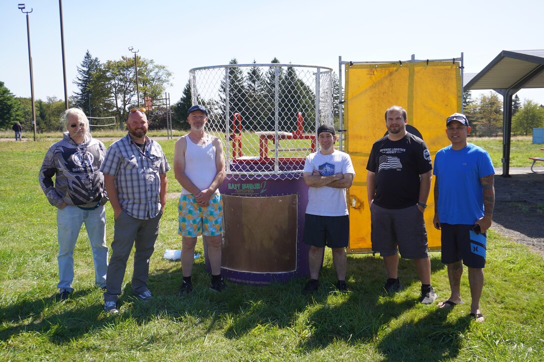 A group of six men stand in front of a dunk tank outside in the grass.