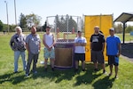 A group of six men stand in front of a dunk tank outside in the grass.