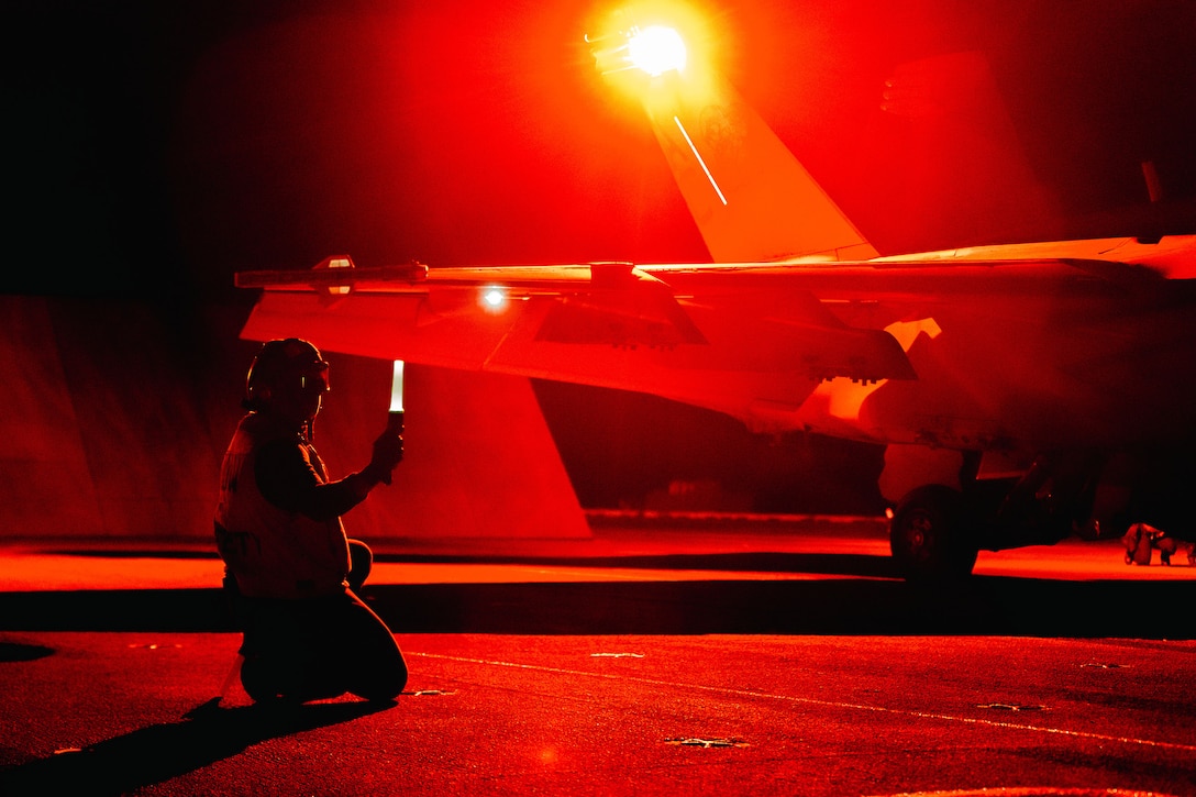 A sailor, illuminated by a red light, kneels to signal a military aircraft aboard the flight deck of a ship at night.