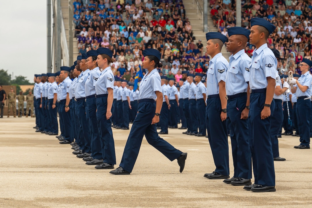 An airman steps forward to join dozens for fellow airman standing in formation during a ceremony as hundreds of spectators watch from a stadium.