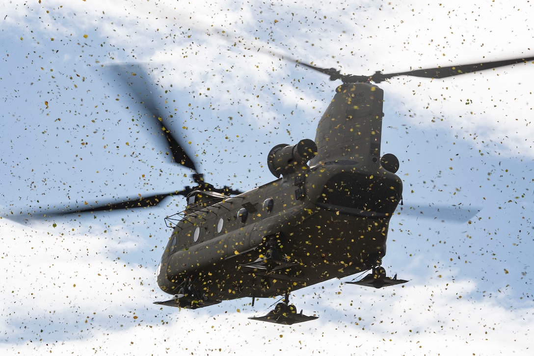 A military helicopter kicks up debris as it hovers above ground during daylight.