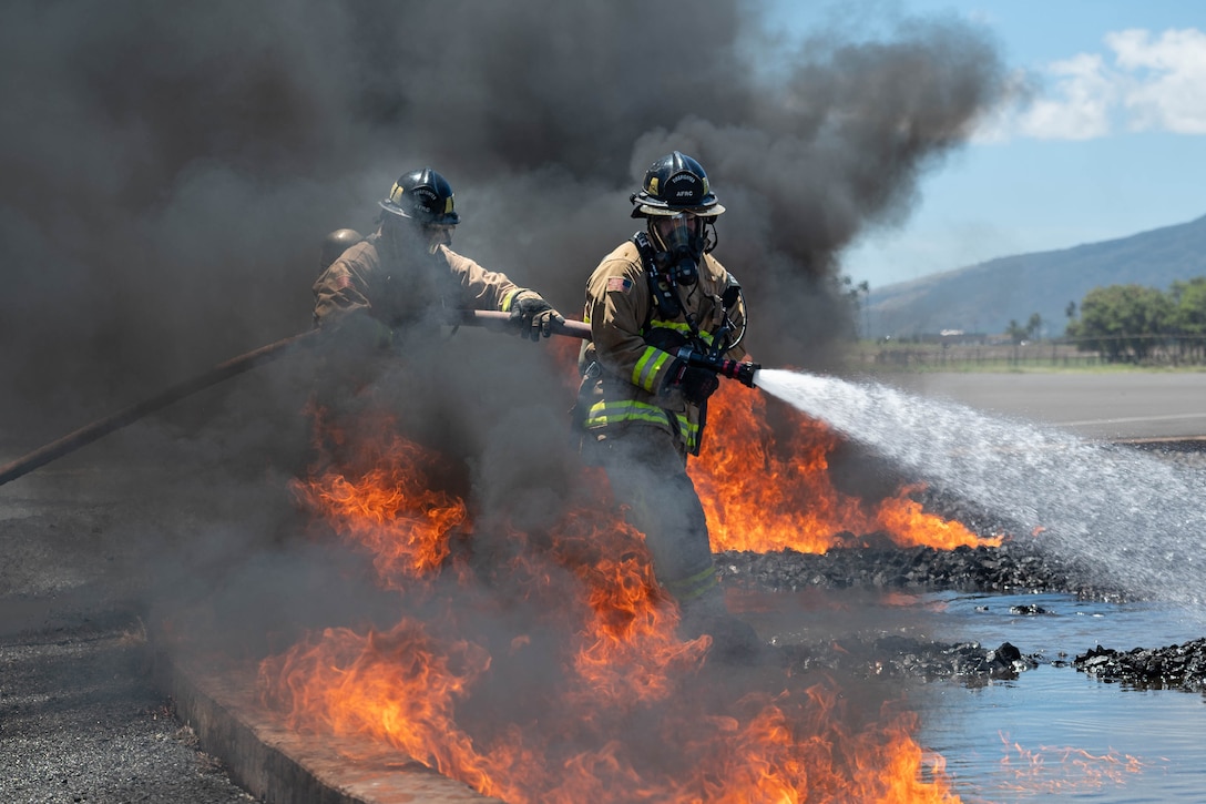 Two airmen firefighters operate a hose to extinguish a jet fuel fire during daylight training as orange flames and dark smoke surround them.