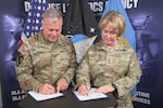 A man and a woman in camo uniforms stand at a table signing documents