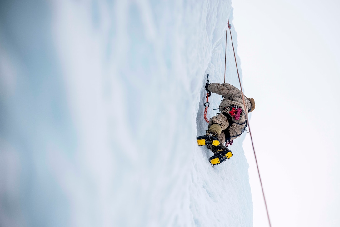 A Marine wearing a harness climbs a snowy mountain as seen from below.