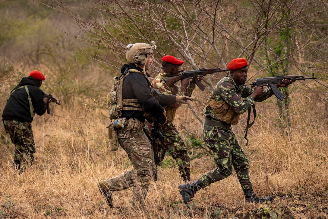 A U.S. soldier and four Mozambican troops, wearing red hats, run through the woods while aiming weapons.