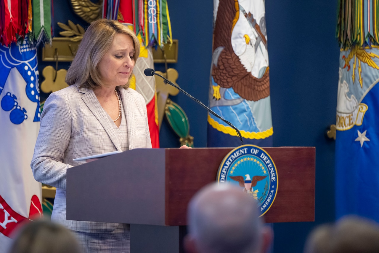 The Deputy Secretary of Defense speaks at a podium in front of various flags and looks down.