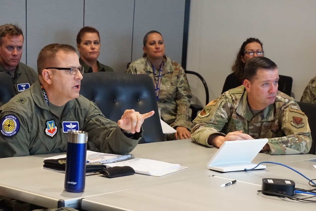 two uniformed U.S. Air Force Airmen sit at conference room table, four individuals are sitting in chairs behind them.