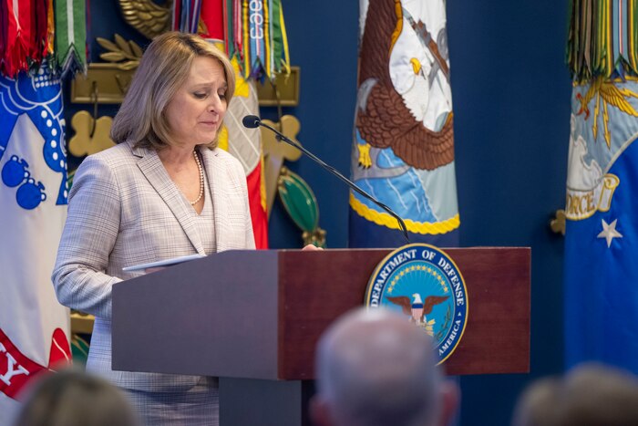The Deputy Secretary of Defense speaks at a podium in front of various flags and looks down.