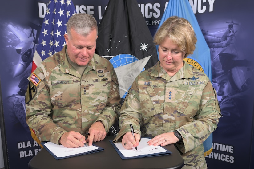 A man and a woman in camo uniforms stand at a table signing documents