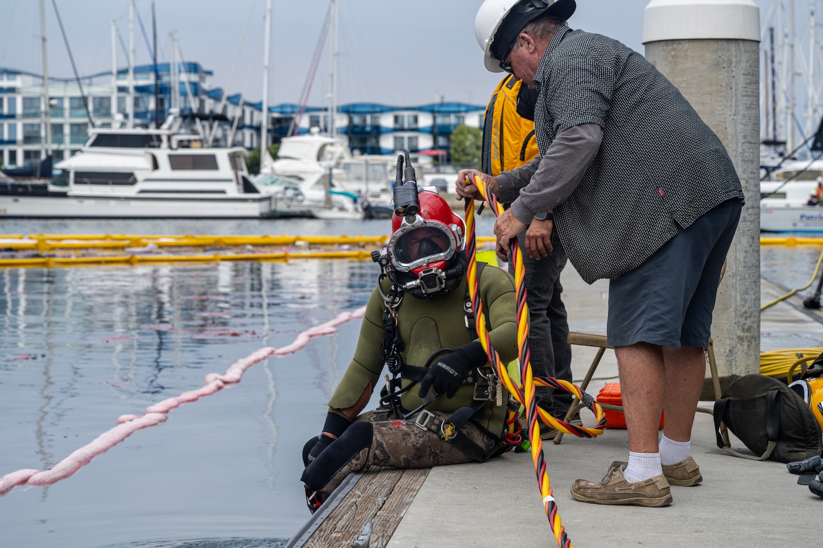 A scuba diver sits on a pier while another man tends his air line.