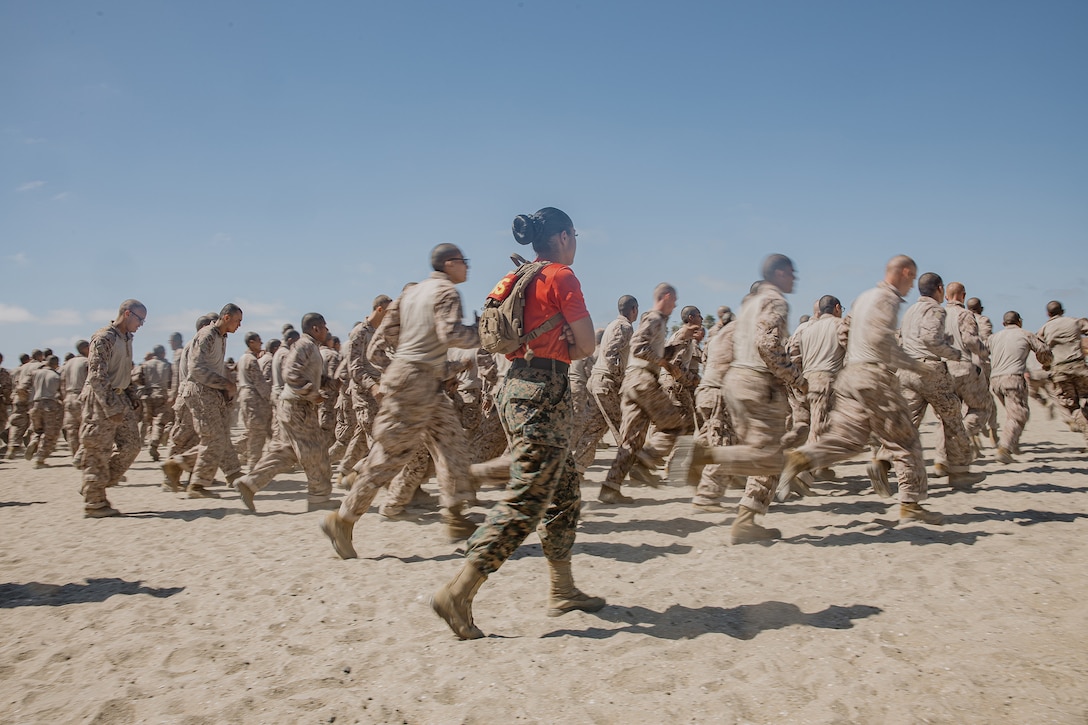 A large group of Marine Corps recruits jog in the sand during daylight. The center service member wears a bright orange shirt.