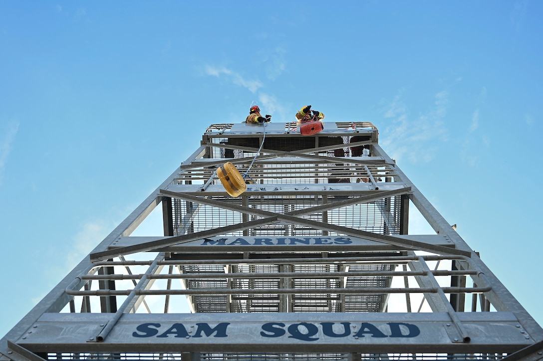 Ground-level photo of airmen hoisting tools up a tower during daylight.