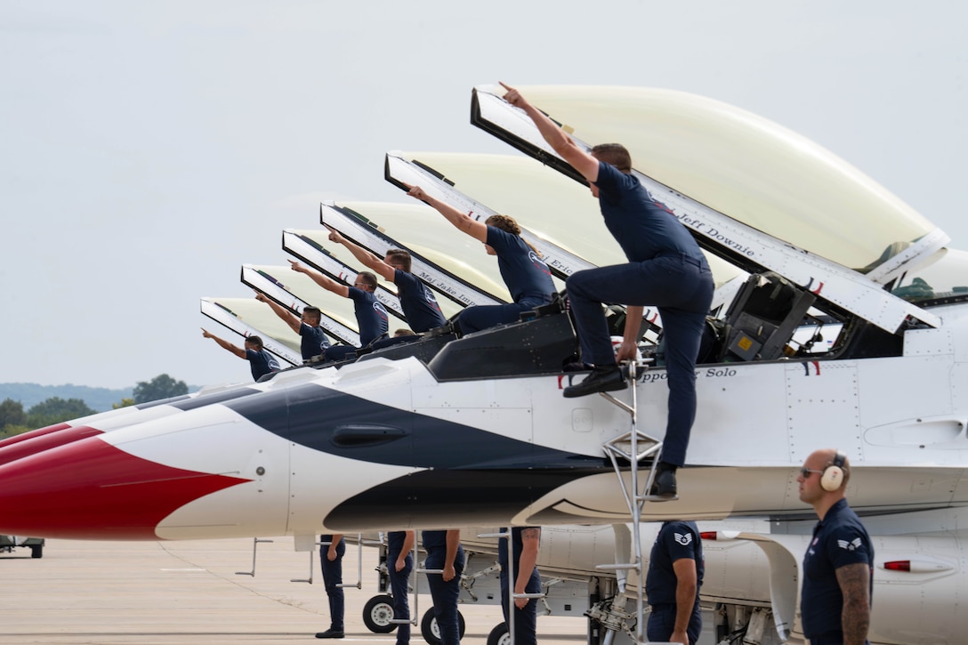 Six airmen on ladders point to the sky while standing next to aircraft with open cockpits as six fellow airmen stand in formation next to the aircraft.