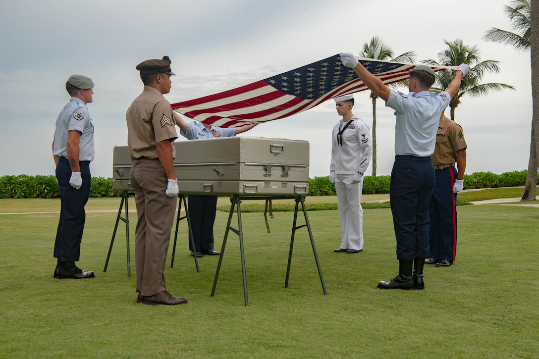 Two service members hold an American flag in the air as fellow service members stand watch near a casket.