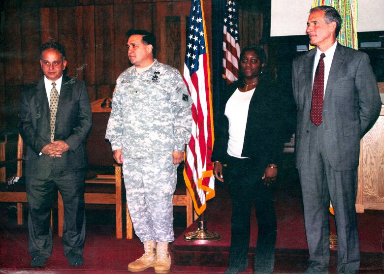 Four people appear on a stage. Three are in business attire and one is wearing an Army uniform