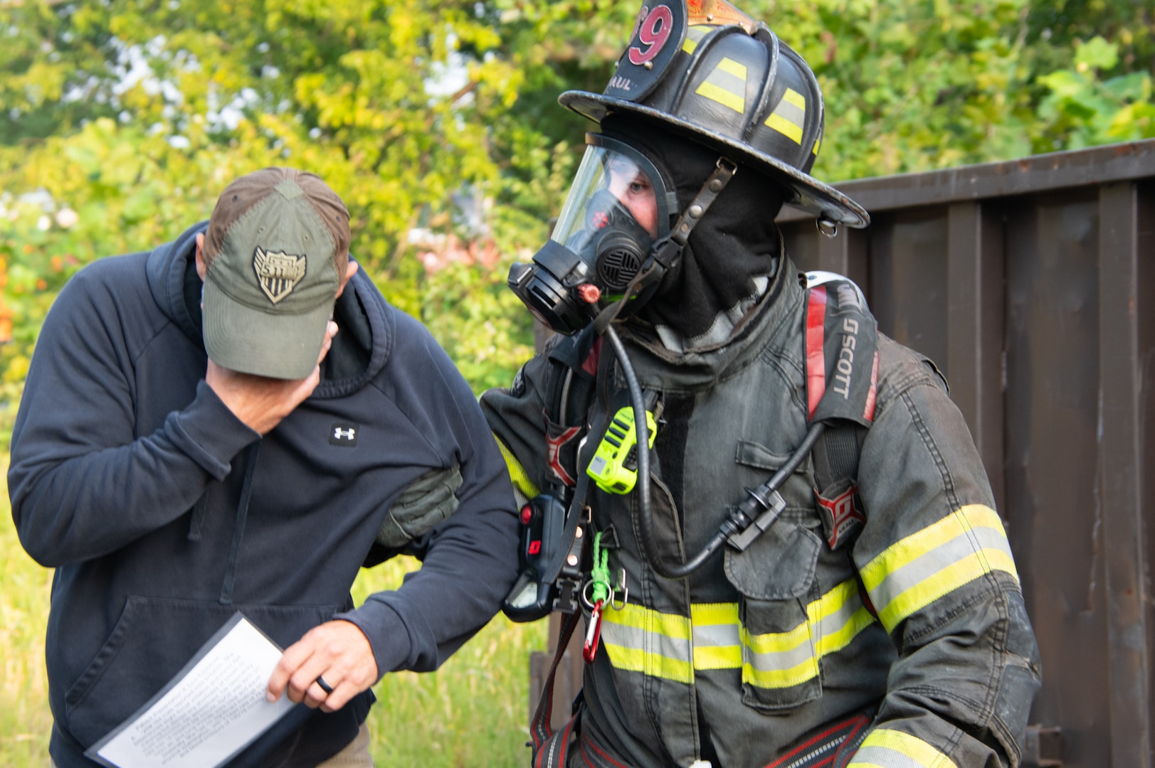 A firefighter in full firefighting equipment guides a man by the arm who is wearing a baseball cap and has his head down with his hand covering his eyes.