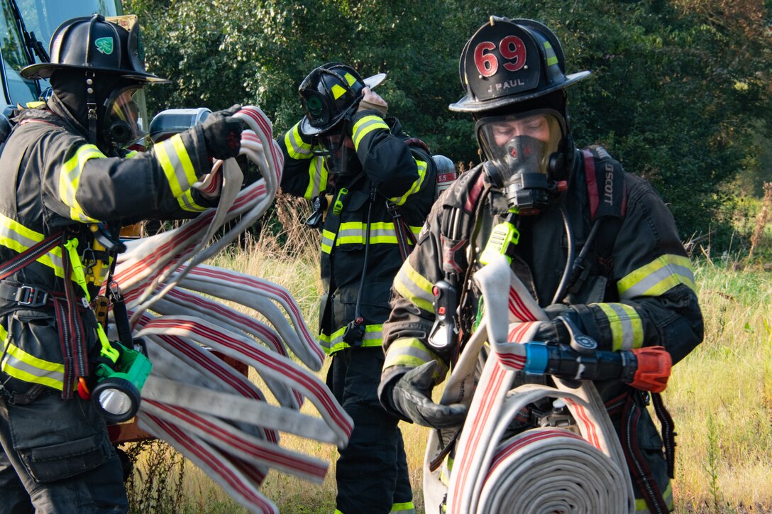 Three firefighters in full firefighting equipment unreel firehoses outside in front of grass and trees.