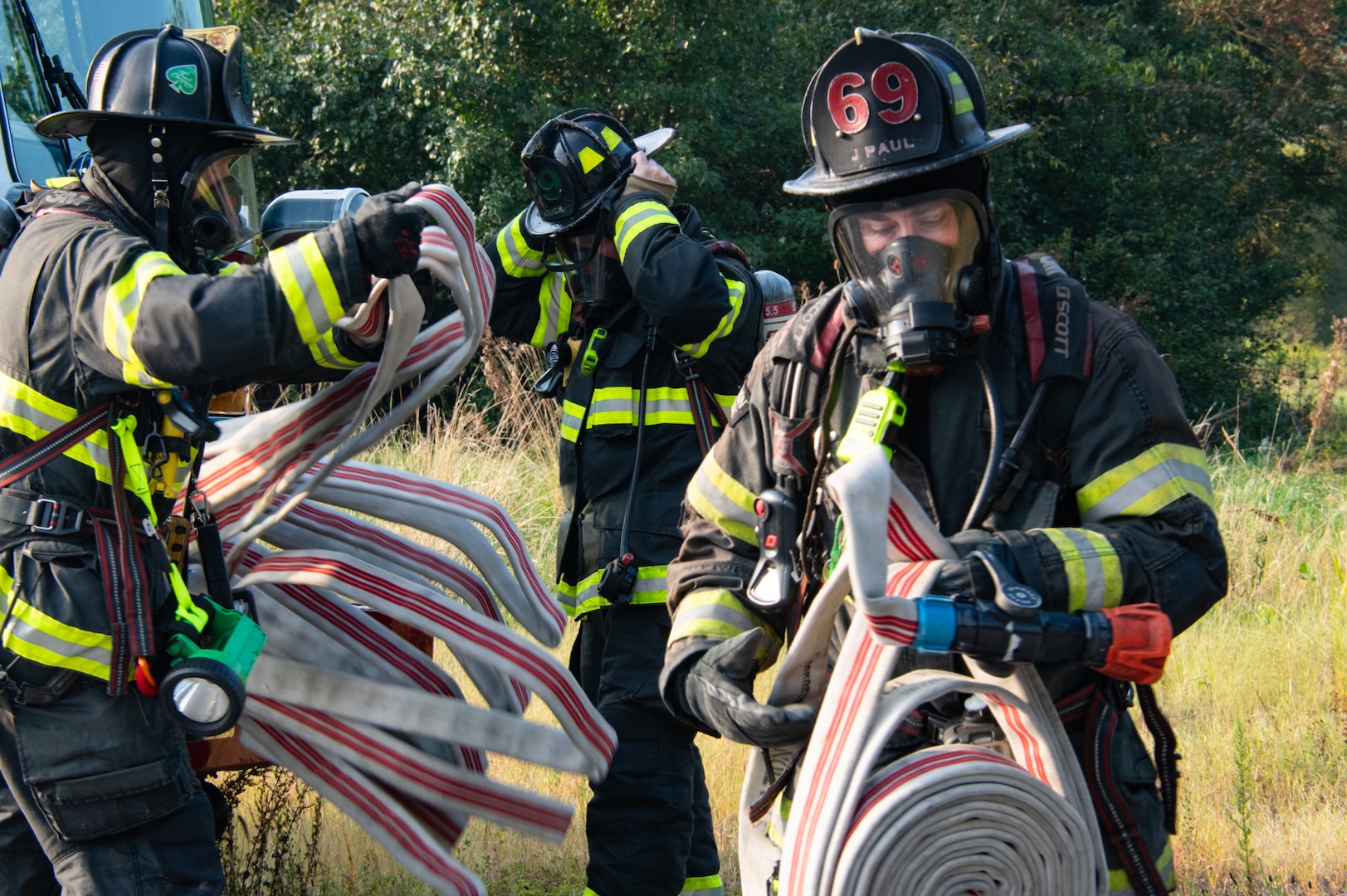 Three firefighters in full firefighting equipment unreel firehoses outside in front of grass and trees.