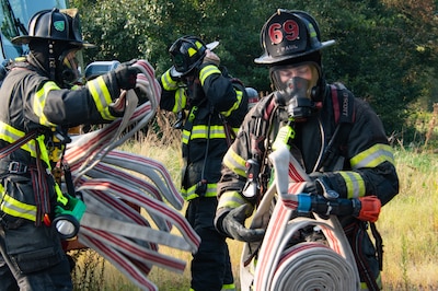 Three firefighters in full firefighting equipment unreel firehoses outside in front of grass and trees.