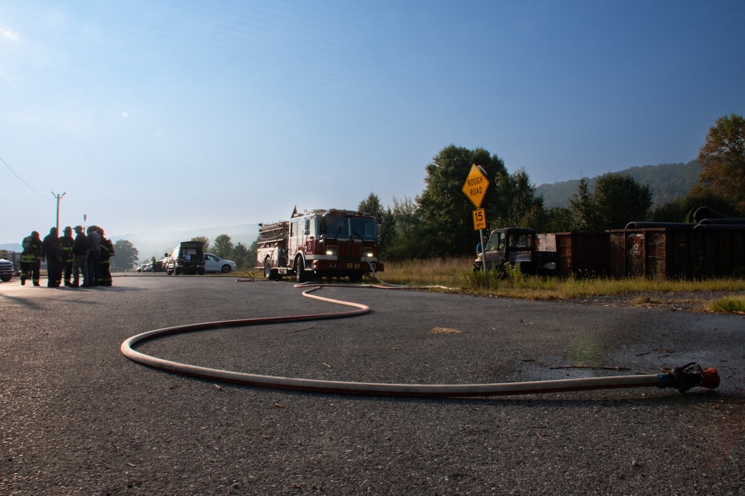 A firetruck with its headlights on is in the background with a firehose extending from it on the road to the foreground with a group of people standing to the left of the firetruck outside on a sunny day with blue skies.