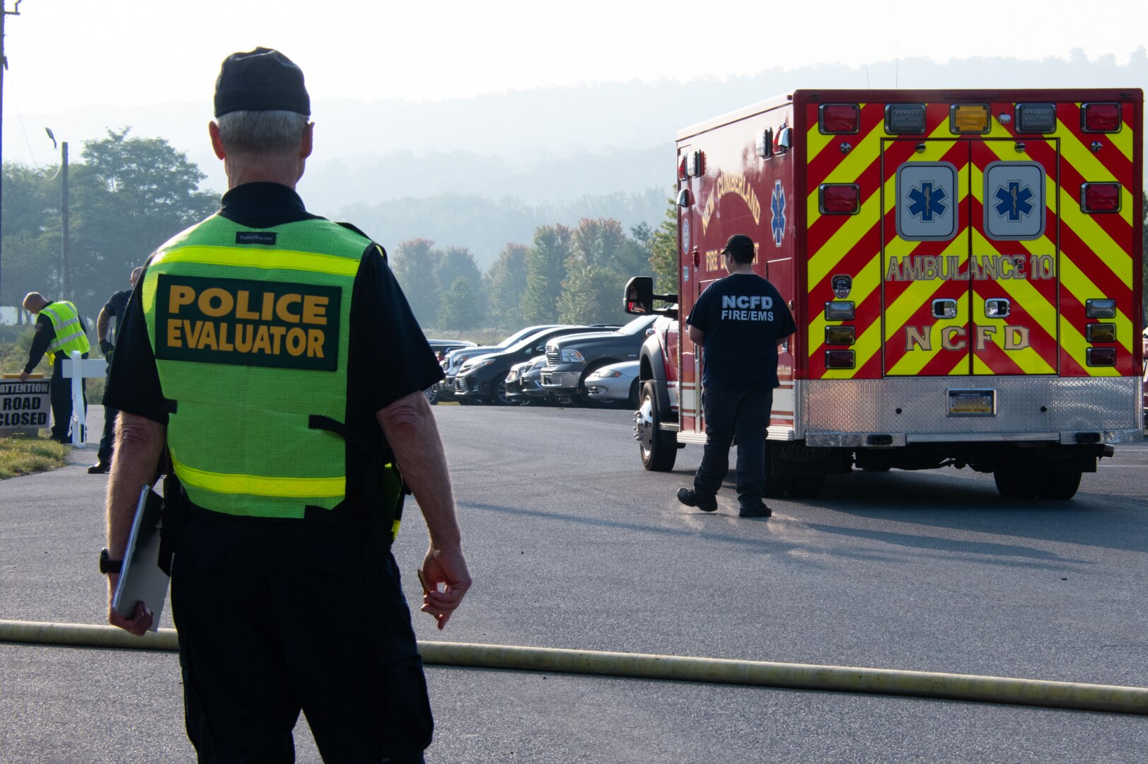 A police officer wearing a yellow safety vest with the words "police evaluator" on it stands in the foreground looking across a road where an ambulance is parked.