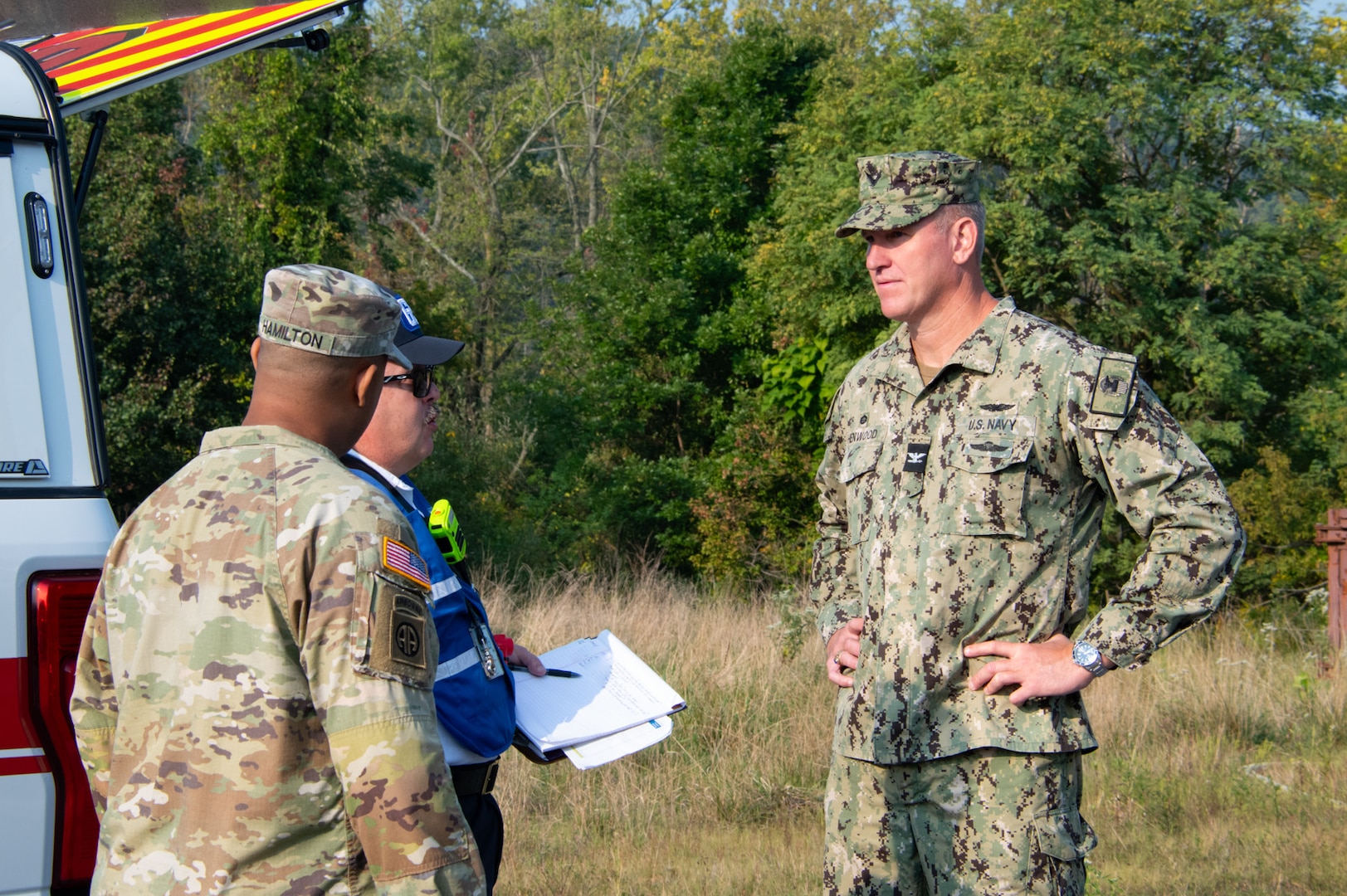 Two men in camouflage uniforms stand outside talking to another man. There are trees and grass in the background.