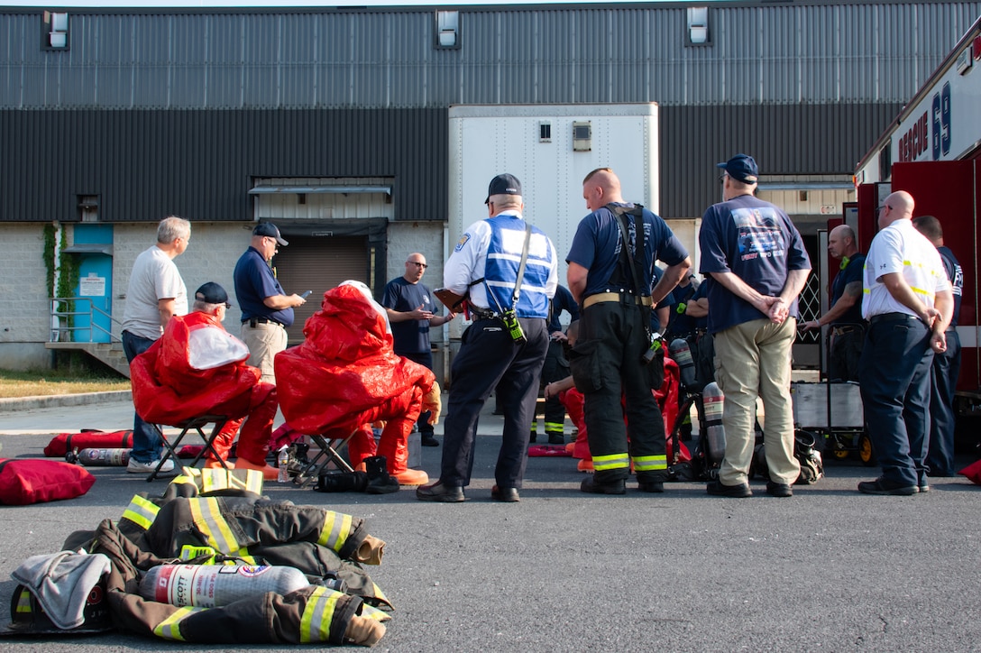 A group of police officers and firefighters stand in a parking lot listening to a man who is speaking