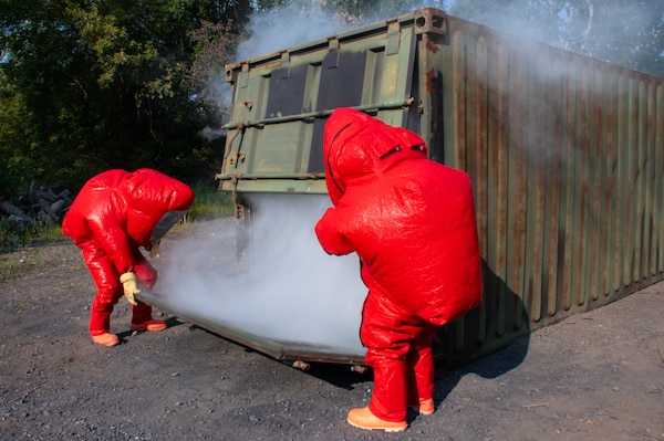Two men in red protective hazardous material suits open a green shipping container with smoke coming out of it outside in a gravel parking lot.
