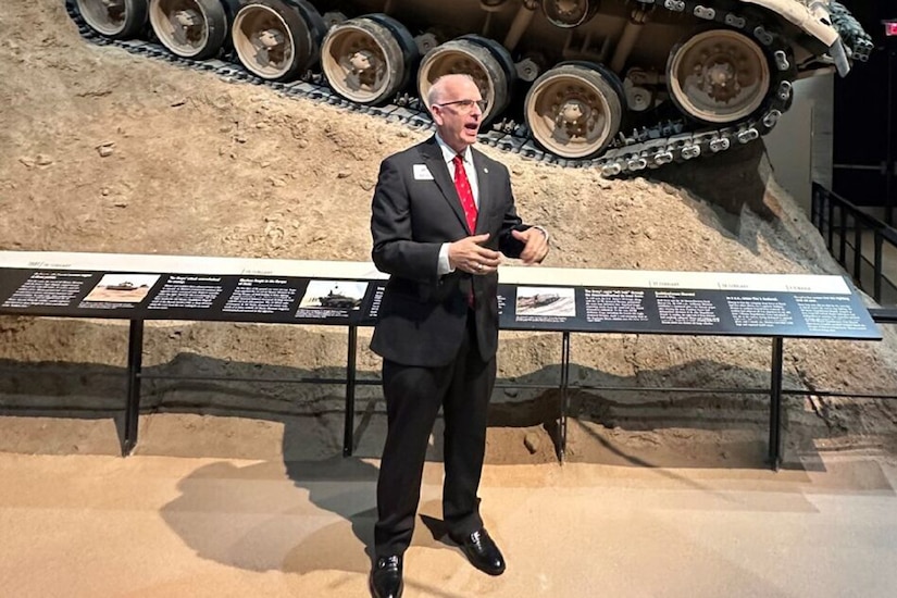 A person in a business suit stands in front of a tank speaks.