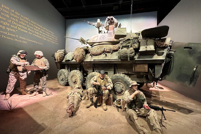 Mannequins dressed as Marines rest next to a tactical vehicle in the desert while the Marine on top looks through binoculars in a museum exhibit.