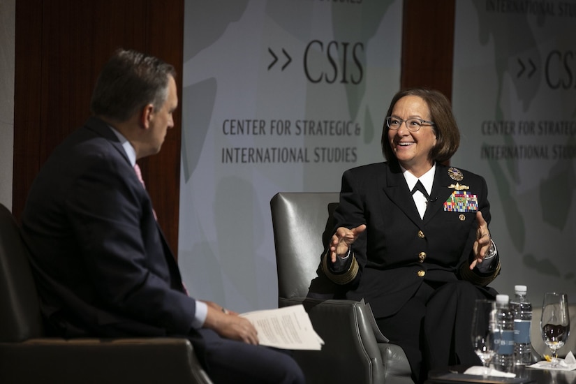 A service member in uniform sits on a stage next to a man in civilian attire.