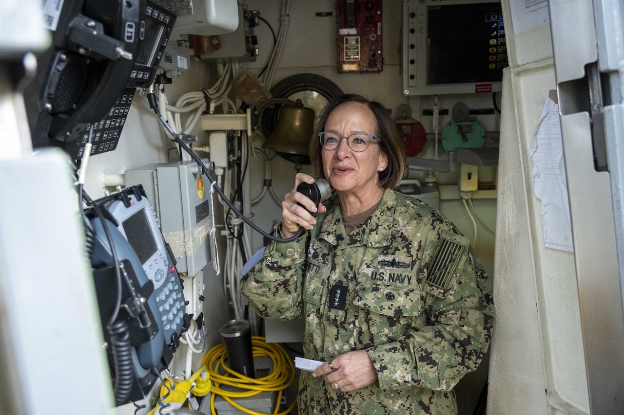 A service member in uniform speaks into a microphone aboard a ship.