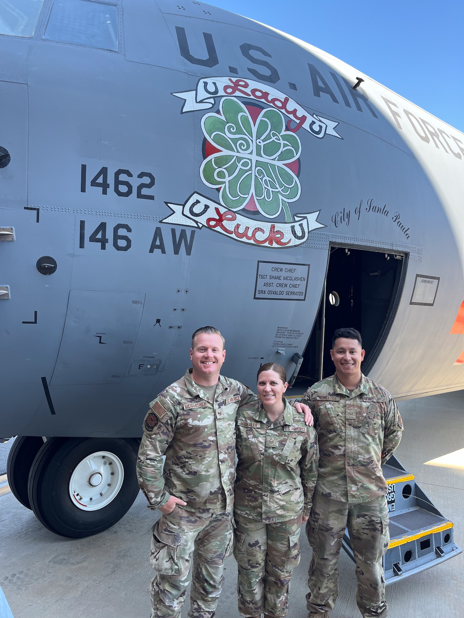 Three Air National Guard Airmen pose in front of the flightdeck door of a C-130J aircraft with nose art that reads "Lady Luck" with a four-leaf clover.